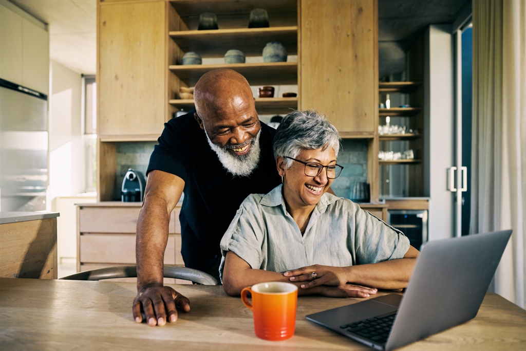 A cheerful older couple sits in a cozy kitchen, smiling as they look at a laptop screen together